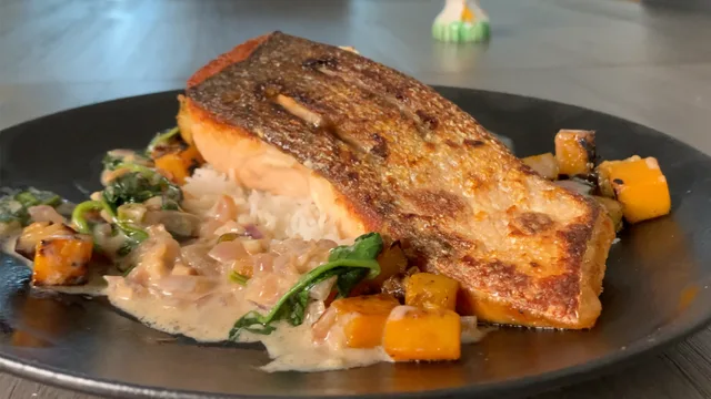 Crispy-skinned salmon fillet on creamy coconut sauce with diced squash and peas on black plate.