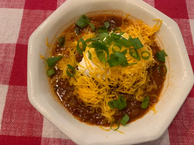 Chicken and bean chili in a white bowl, topped with shredded cheddar, a spoonful of sour cream and sliced green onions, on a red gingham tablecloth.