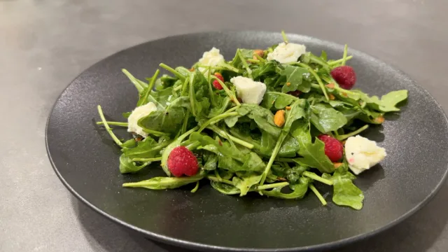 Arugula salad with fresh raspberries, creamy goat cheese, and colorful vegetables on a dark plate.