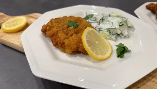 Breaded and fried wiener schnitzel served on white plate with lemon slices and creamy cucumber salad.