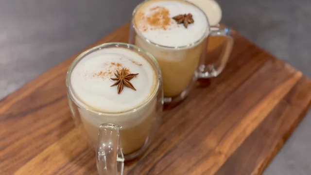 Two mugs of dirty chai latte with creamy foam tops, star anise garnish, and cinnamon dust on a wooden board.
