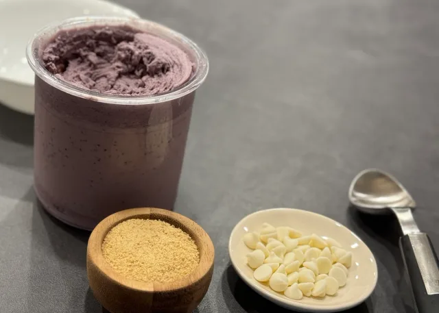 Blueberry cheesecake ice cream in a clear container with graham cracker crumbs and white chocolate chips beside it.