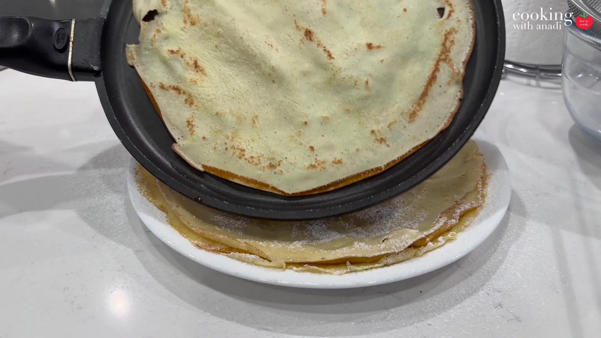 A stack of thin crepes on a plate, one still in the pan