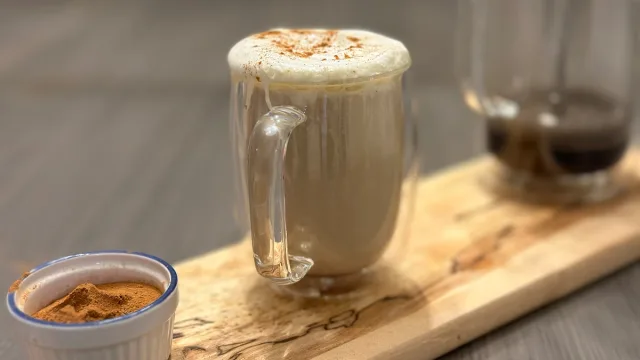 Homemade eggnog latte with cinnamon foam in a glass mug, cinnamon powder in small bowl, on wooden board.