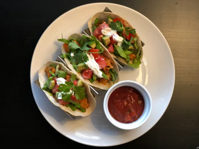 Three crispy fish tacos topped with fresh cilantro, diced tomatoes, sour cream, and green onions, served with salsa on the side.