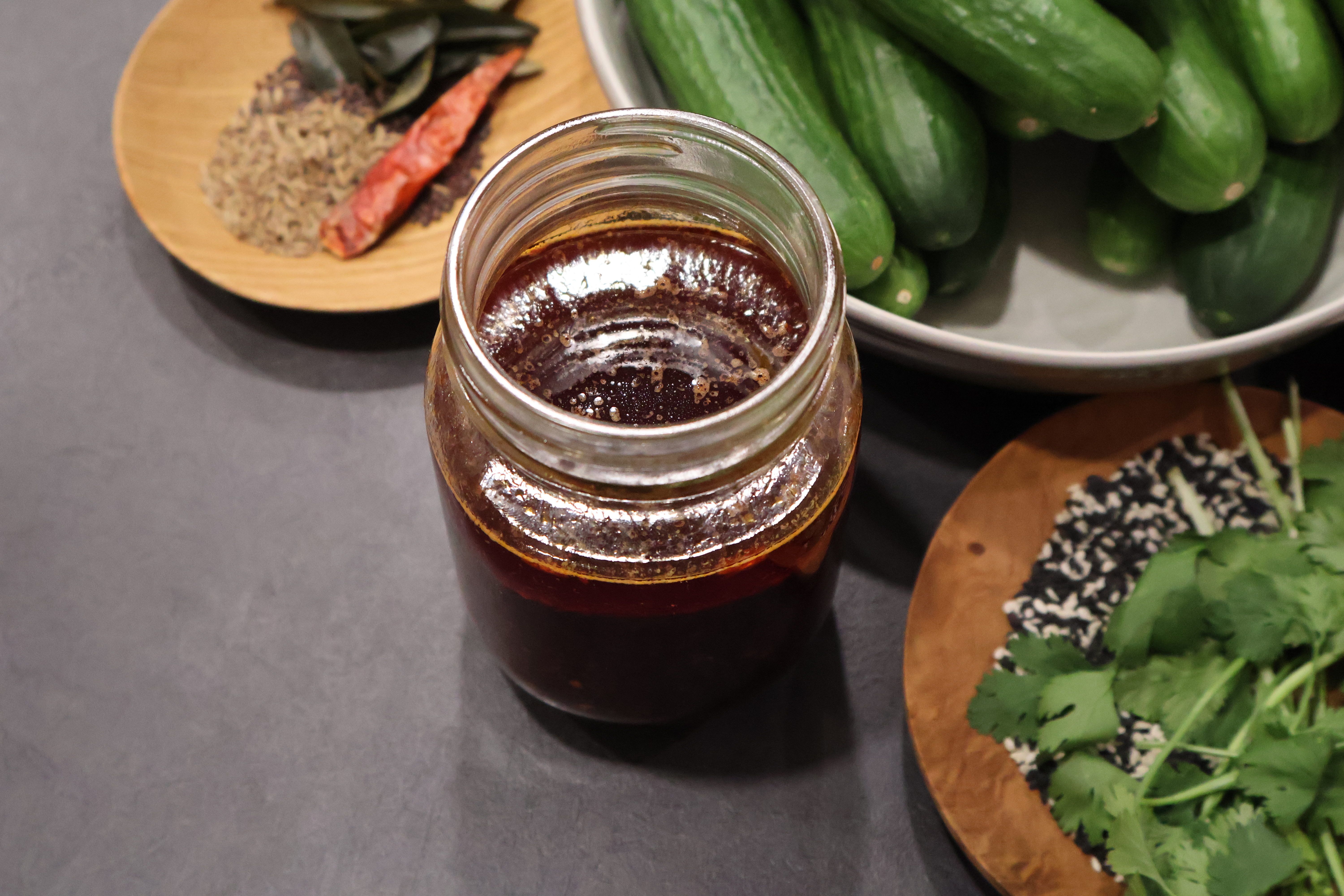A glass mason jar of dark soy sesame dressing with chili on a slate surface, surrounded by whole cucumbers, fresh cilantro, black and white sesame seeds, and a dried red chili.