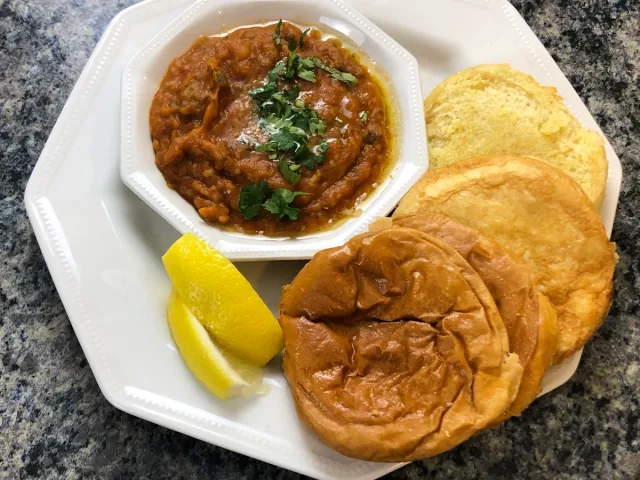 Bombay pav bhaji plated with spiced vegetable curry, toasted bread rolls, and lemon wedge on white dish.