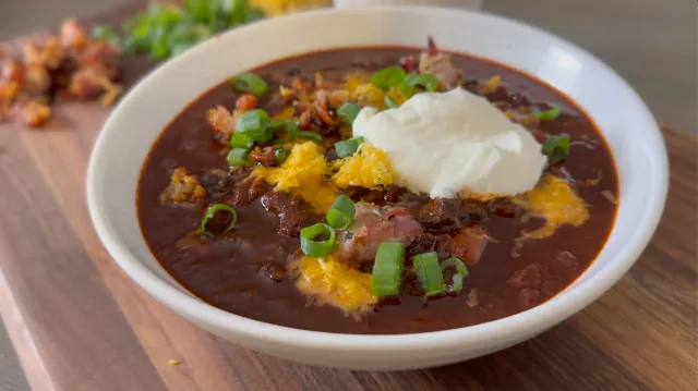 Bowl of dark red chili topped with cornbread crumbles, sour cream, and sliced green onions.