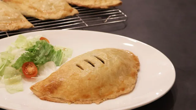 Golden-brown handheld tourtiere on white plate with lettuce and tomato garnish, cooling rack with baked pastries in background.