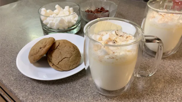 White hot chocolate topped with cinnamon in glass mugs beside ginger cookies, marshmallows, and ingredients.
