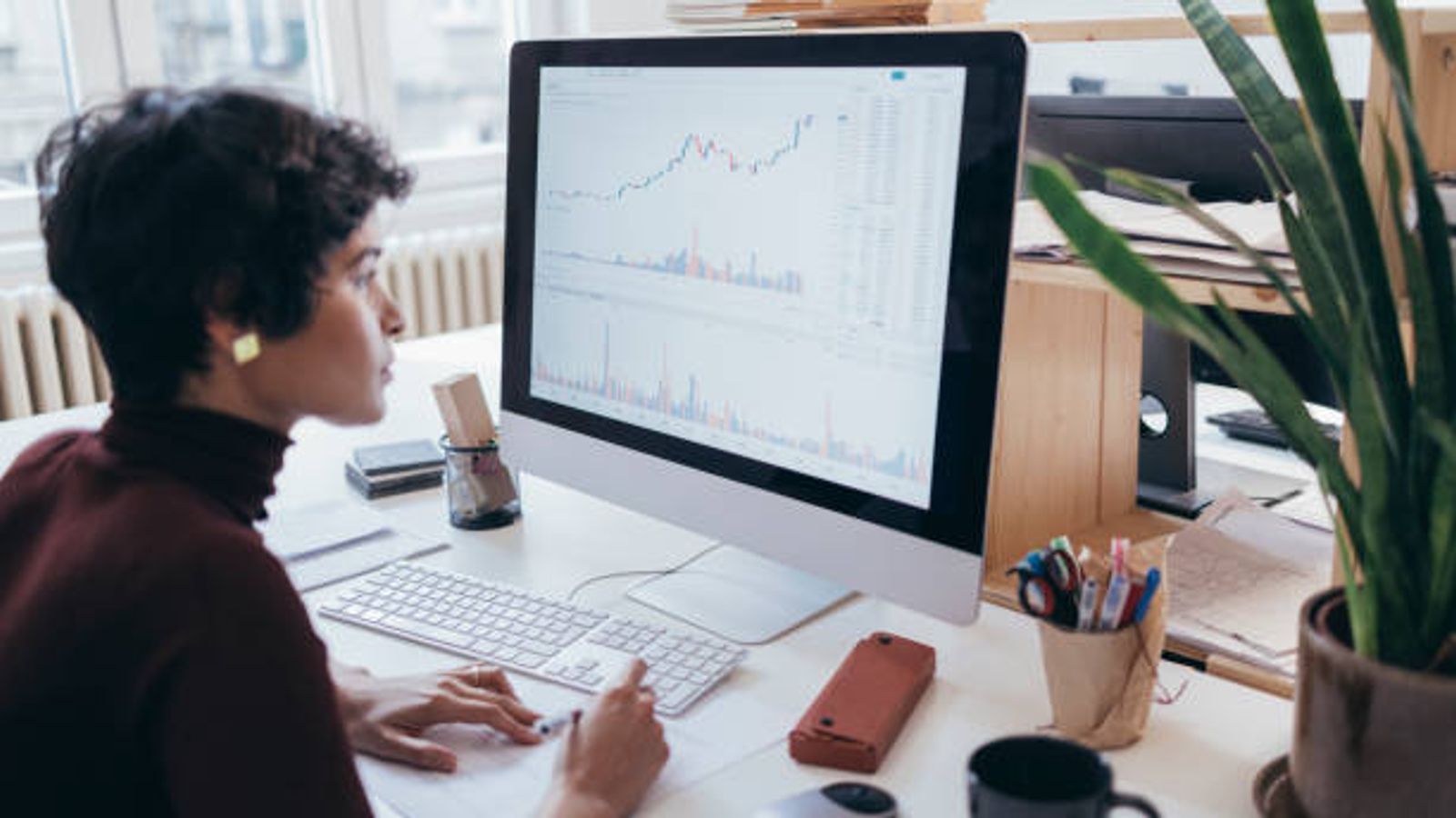 A person sits at a desk working on a computer displaying charts, graphs, and SEO metrics like click-through rate (CTR), taking notes on paper. The workspace is bright with natural light, featuring office supplies, a plant, and a coffee mug.