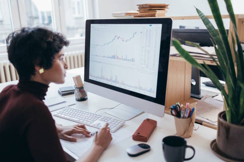 A person sits at a desk working on a computer displaying charts, graphs, and SEO metrics like click-through rate (CTR), taking notes on paper. The workspace is bright with natural light, featuring office supplies, a plant, and a coffee mug.