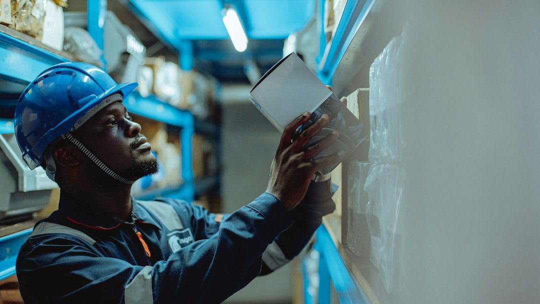 A worker in a hard hat and coveralls checking a box on a stockroom shelf.