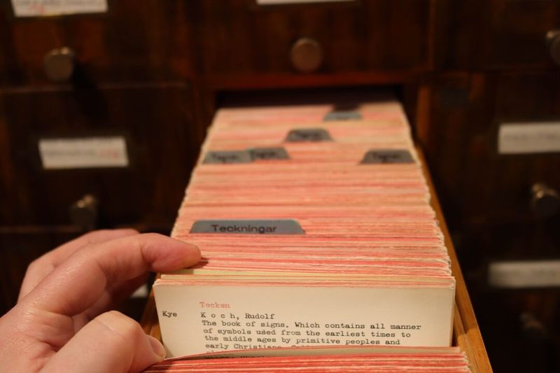 A hand lifting an index card from a library card catalogue drawer, with the surrounding cards and drawer labels visible.
