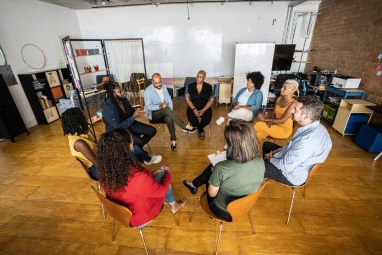 A group of nine people is seated in a circle in a brightly lit room with wooden floors. They are engaged in a discussion on competitive intelligence, with some holding papers or notebooks. Shelving, a whiteboard, and various office supplies are visible in the background.