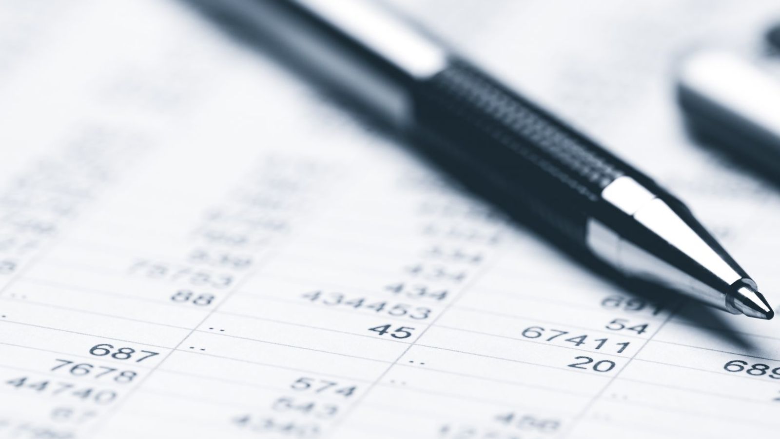 A black and silver pen rests on a sheet filled with numbers and data, next to a partially visible calculator. The sheet appears to be a financial or accounting document with columns of numbers and figures, adhering closely to pricing principles.