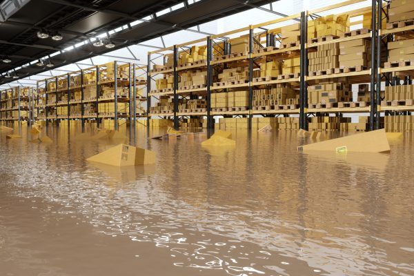 A warehouse with shelves stacked with numerous cardboard boxes. The floor is heavily flooded, causing some boxes to float in the water. The shelves extend to the ceiling, and the area has ample lighting from overhead fixtures-proof that luck favors the prepared.