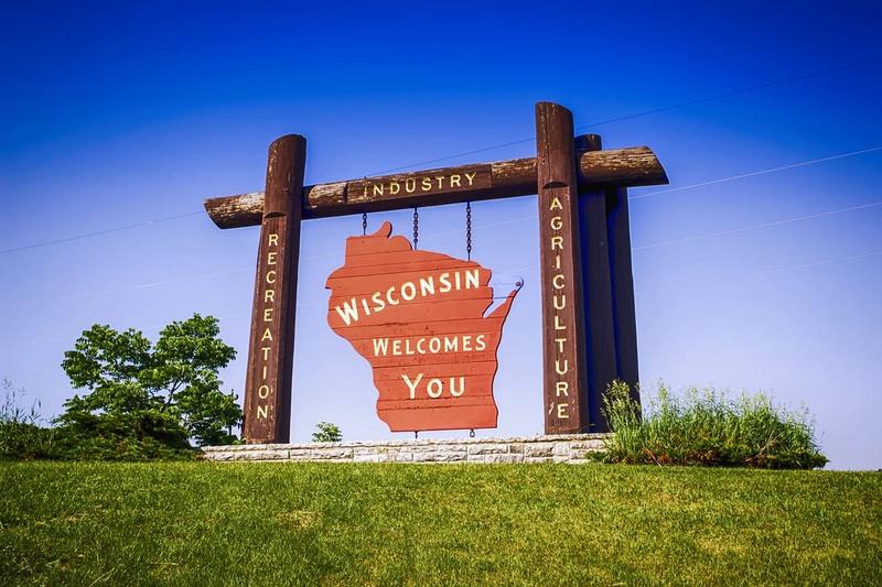 Large wooden sign in the shape of Wisconsin with text reading "Wisconsin Welcomes You," standing on a grassy area against a clear blue sky. The sign includes additional vertical text: "Recreation," "Industry," and "Agriculture" on wooden posts, highlighting the state's offerings with marketing agency precision.
