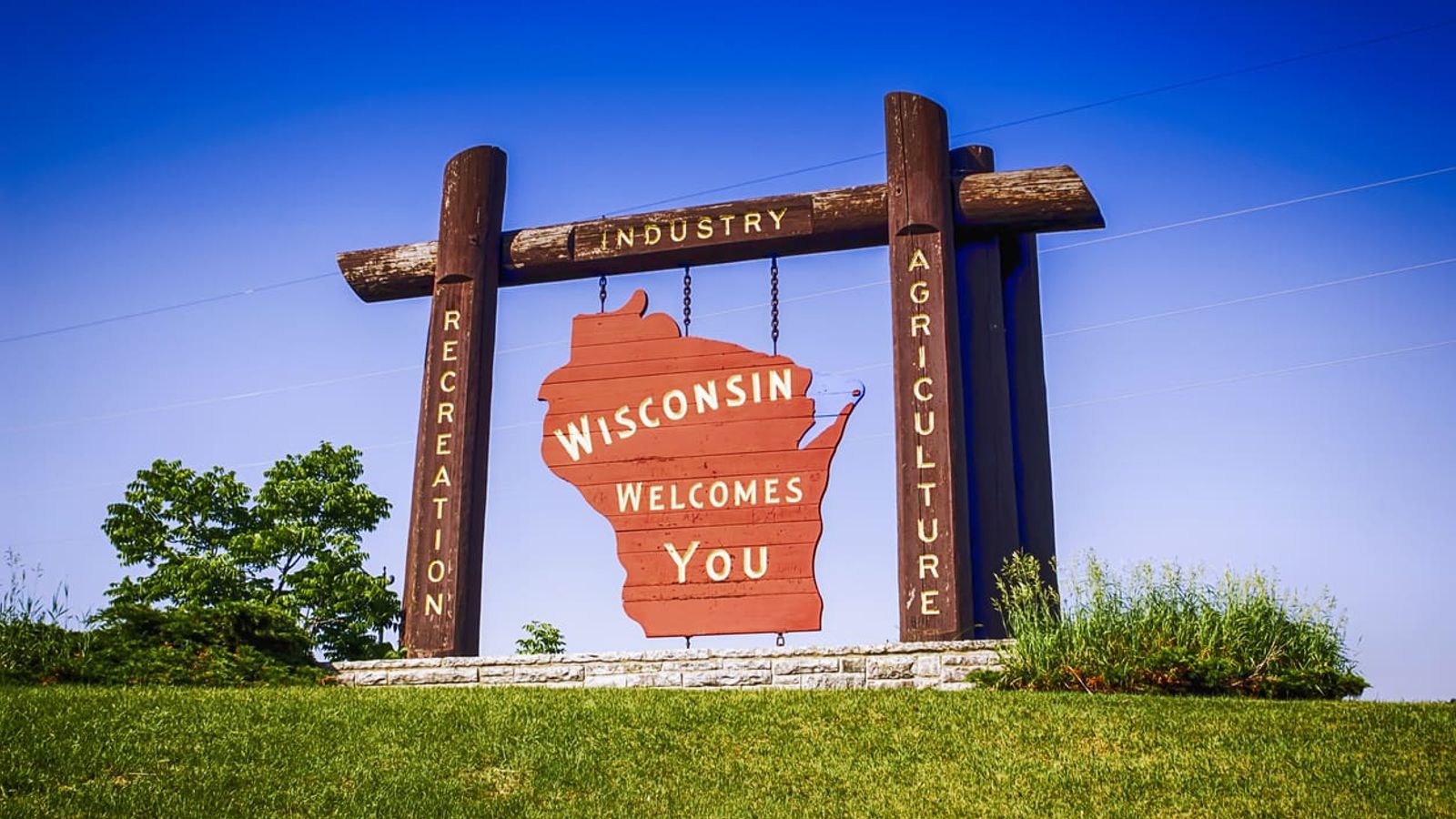 Large wooden sign in the shape of Wisconsin with text reading "Wisconsin Welcomes You," standing on a grassy area against a clear blue sky. The sign includes additional vertical text: "Recreation," "Industry," and "Agriculture" on wooden posts, highlighting the state's offerings with marketing agency precision.