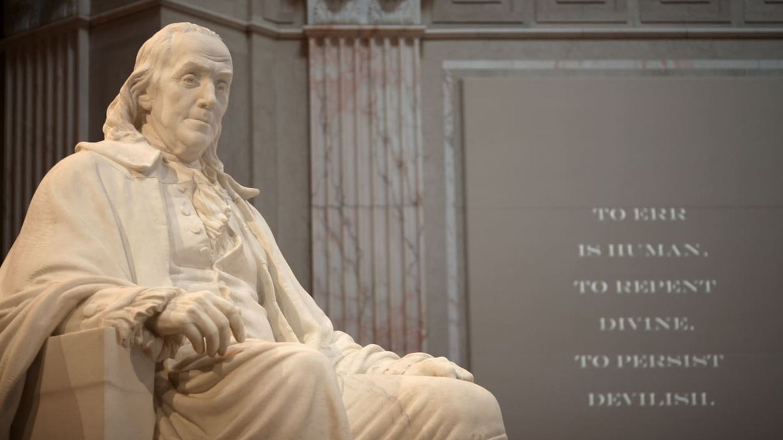 A marble statue of Benjamin Franklin seated and wearing historical attire is prominently displayed indoors. To the right, a wall features the inscription: "To err is human, to repent divine, to persist devilish." The setting is grand with ornate columns in the background, a testament to "Diligence is the Mother of Good Luck.