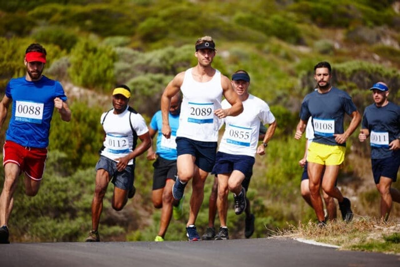 A group of runners wearing numbered bibs participates in an outdoor race on a road surrounded by greenery. The runners, dressed in athletic attire, are captured mid-stride with determined expressions, embodying the saying "Diligence is the Mother of Good Luck." The sky is clear, and the overall scene is vibrant and dynamic.