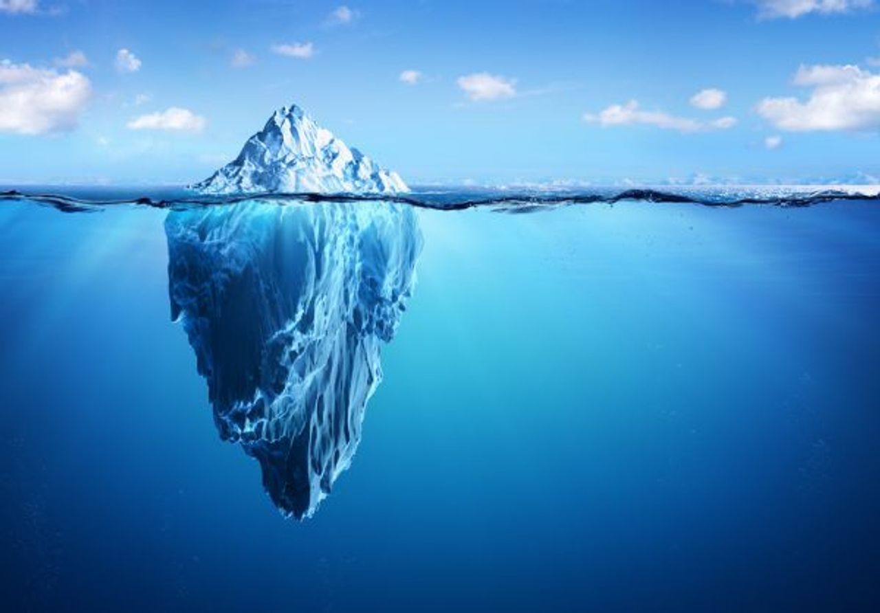 A massive iceberg floats in a blue ocean under a clear sky. About one-ninth of the iceberg is visible above the water, while the vast submerged part is seen beneath the surface, showcasing dark blue and icy textures below the waterline. Truly, luck favors the prepared who navigate these treacherous waters.