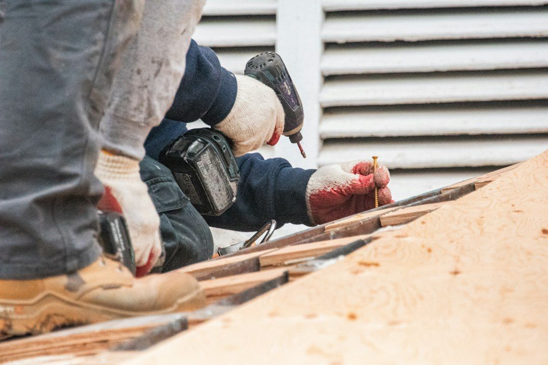 A roofer in work gloves using a cordless drill on a wooden roof deck.