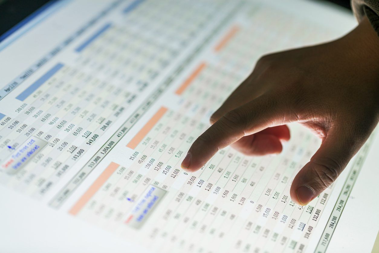 A hand points at numbers on a financial spreadsheet displayed on a screen. The spreadsheet contains various rows and columns with numerical data, highlighted cells, and different colored text, indicating a detailed analysis of the marketing budget.