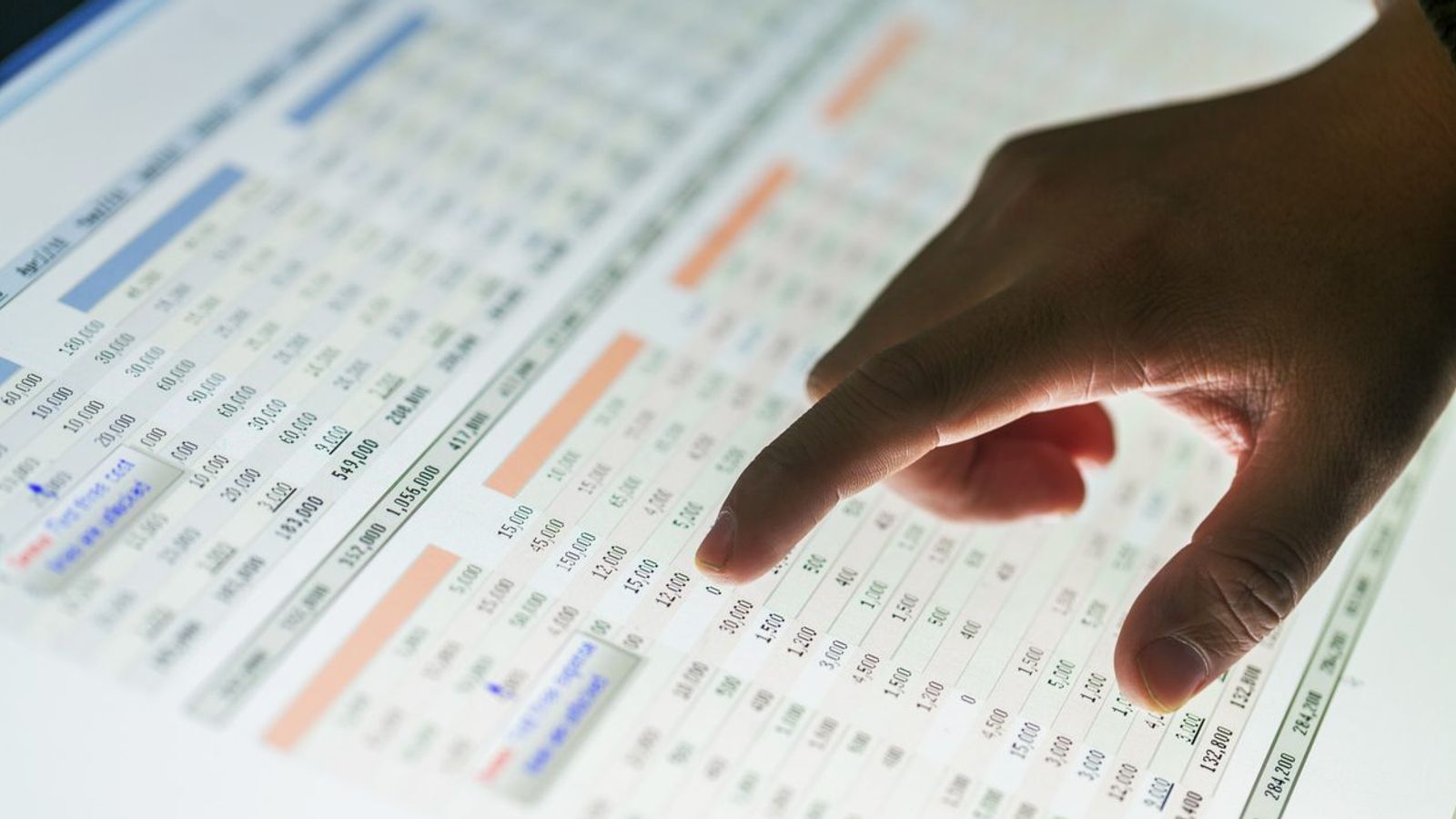 A hand points at numbers on a financial spreadsheet displayed on a screen. The spreadsheet contains various rows and columns with numerical data, highlighted cells, and different colored text, indicating a detailed analysis of the marketing budget.