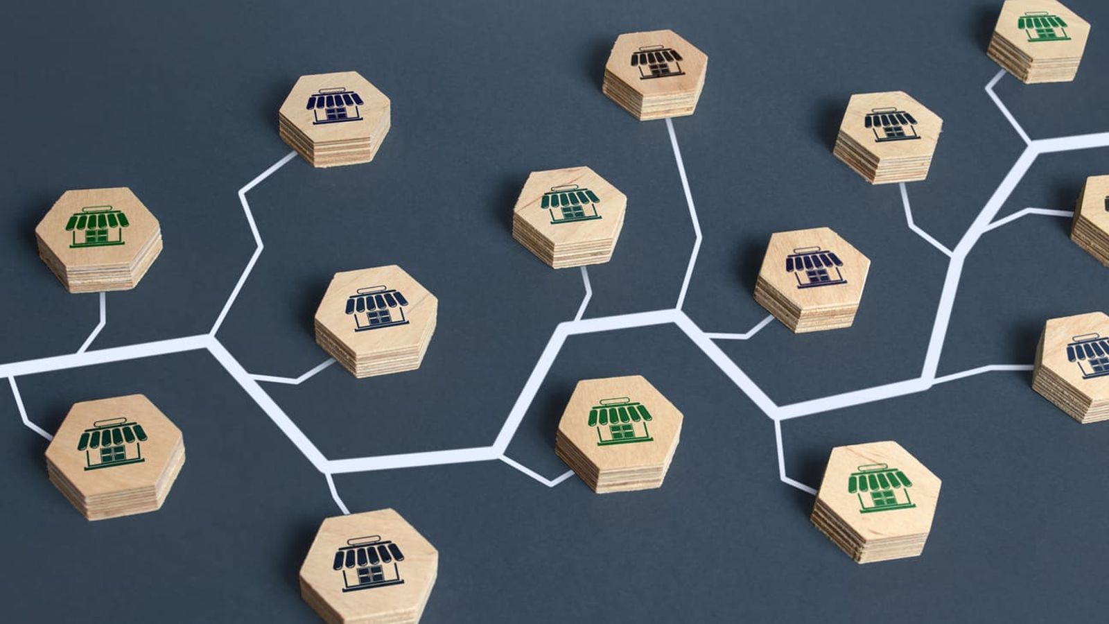 An abstract representation of a franchise network or organizational structure using wooden hexagons with store icons interconnected by white lines on a dark background, illustrating the importance of franchise SEO. The hexagons feature blue, green, and black storefront symbols.