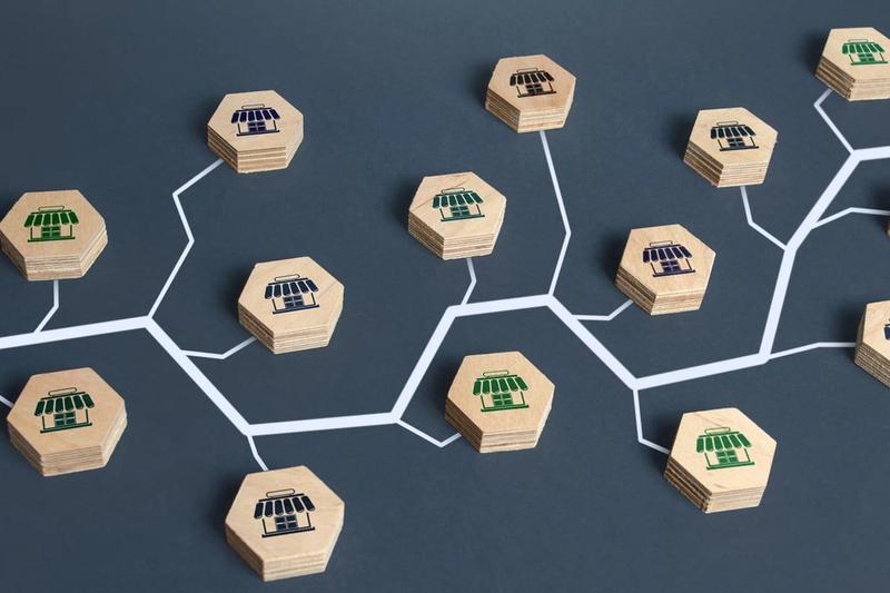 An abstract representation of a franchise network or organizational structure using wooden hexagons with store icons interconnected by white lines on a dark background, illustrating the importance of franchise SEO. The hexagons feature blue, green, and black storefront symbols.