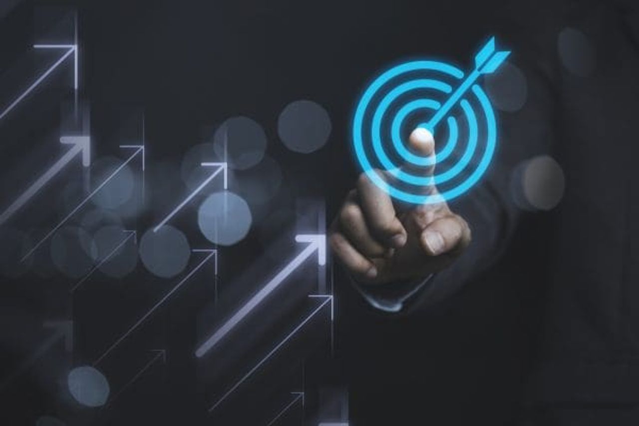 A person in a dark suit points at a glowing blue target symbol on a transparent screen. Several translucent arrows pointing upward can be seen on the left side, symbolizing growth and progress, reflecting the optimization of their marketing budget.