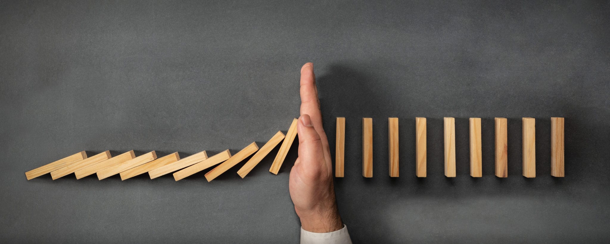 A hand prevents a falling line of wooden dominoes from continuing its chain reaction, symbolizing the precise interventions of a top marketing agency in Madison, WI. The dominoes on the left are tipped over, while those on the right remain standing upright against a plain, dark surface.