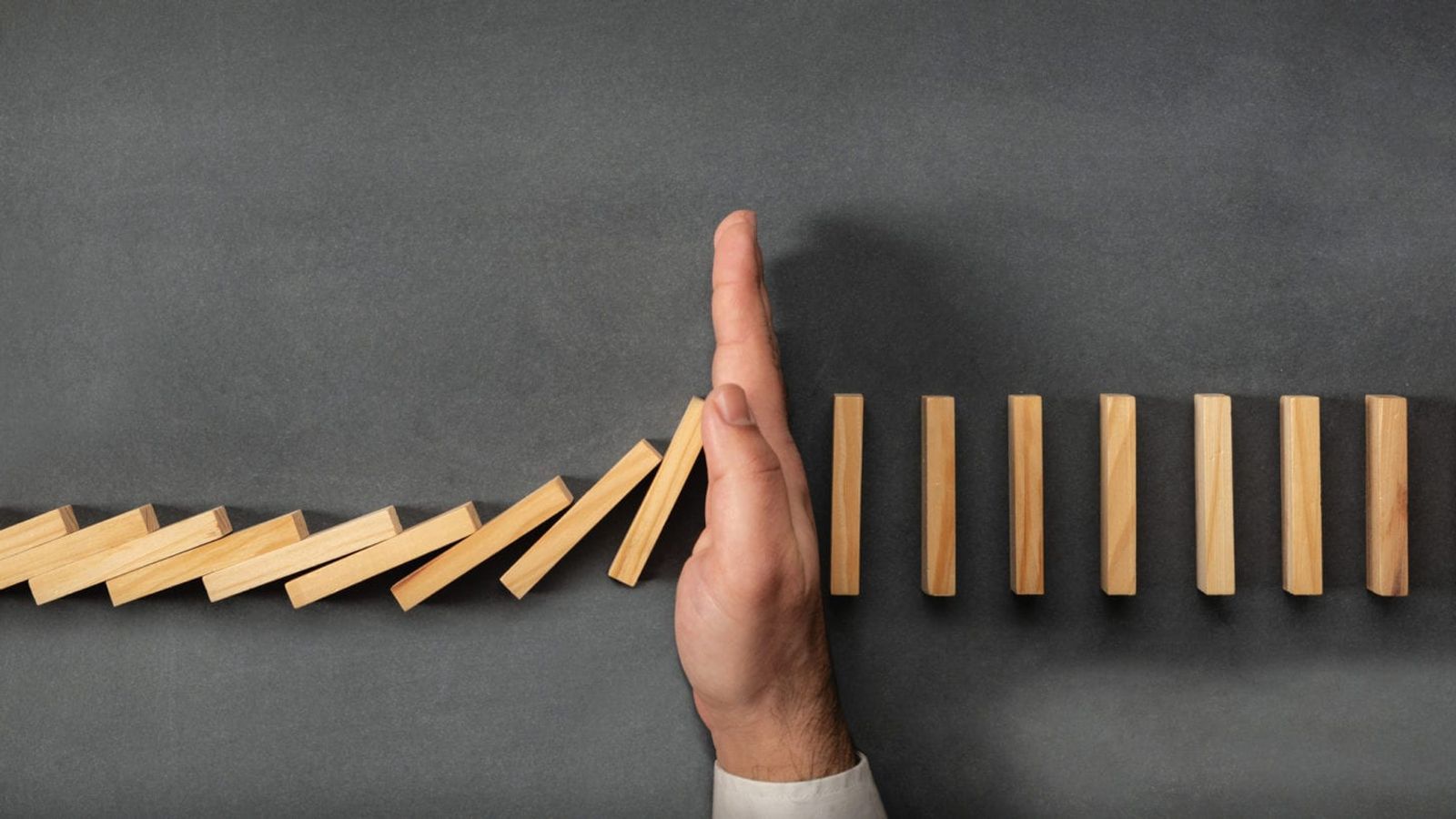 A hand prevents a falling line of wooden dominoes from continuing its chain reaction, symbolizing the precise interventions of a top marketing agency in Madison, WI. The dominoes on the left are tipped over, while those on the right remain standing upright against a plain, dark surface.