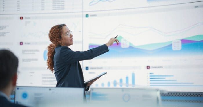 A person with curly hair, wearing glasses and business attire, points to a large digital screen displaying charts and graphs. They hold a tablet in one hand. The background shows multiple digital screens with data and visualizations, emphasizing competitive intelligence.
