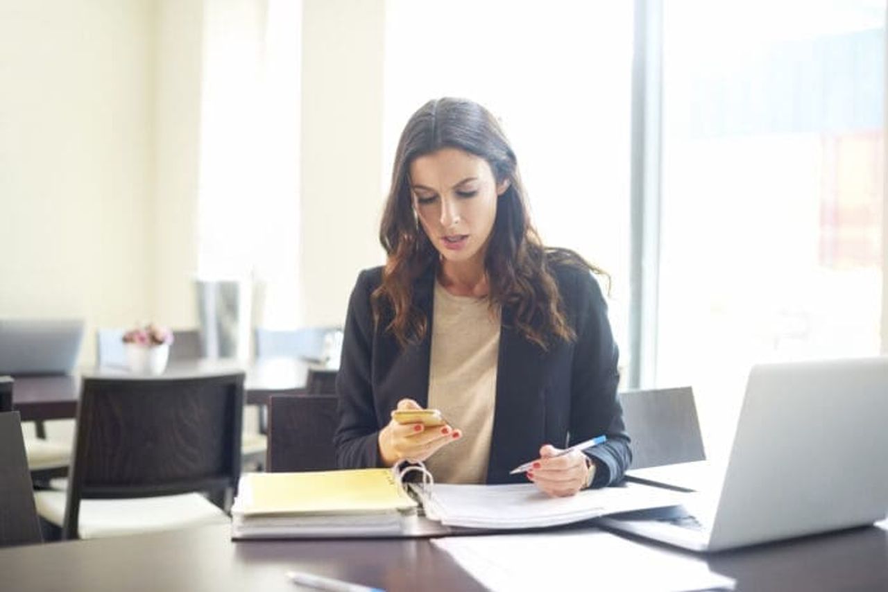A woman sitting at a desk in a bright room, holding a smartphone in her right hand and a pen in her left, exemplifies the saying "Diligence is the Mother of Good Luck" as she looks at a large binder with papers. A laptop is open on the desk beside her.
