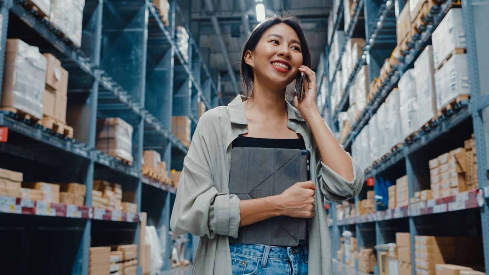 A woman stands in a large warehouse aisle, smiling while talking on a smartphone. She is holding a gray binder and wearing a light green jacket over a black top and jeans. The shelves behind her are filled with various packed boxes, embodying the efficient integration of technology in small business operations.