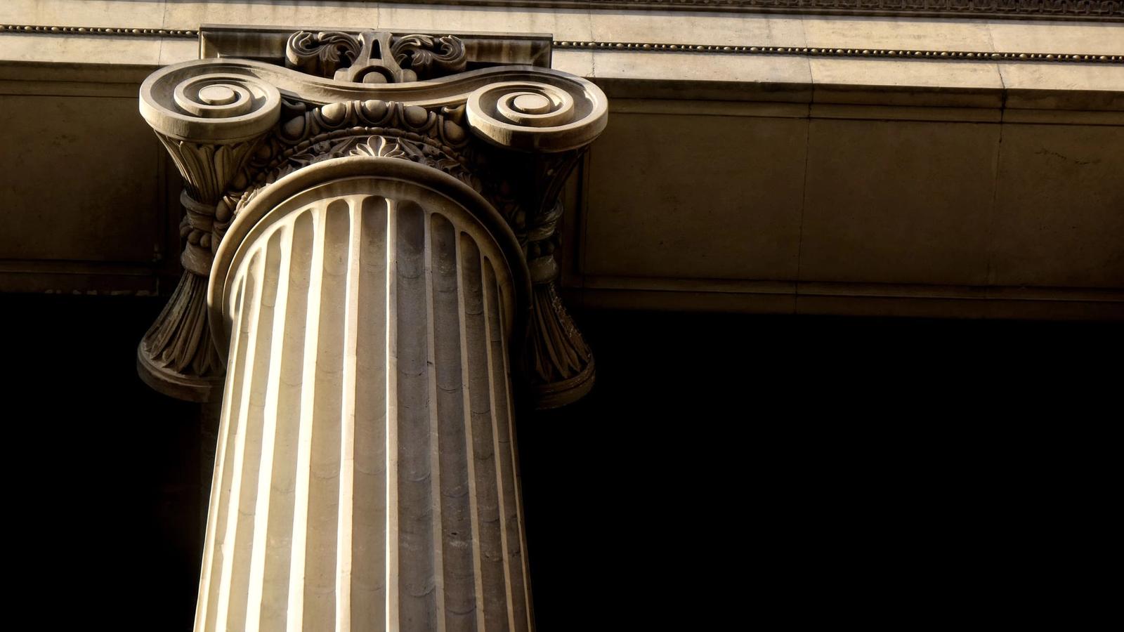 A close-up of a Corinthian column with intricate carvings and decorative scrolls, resembling the structure of a pillar page. The image highlights the column’s detailed capital and fluted shaft set against a dark background, capturing classical architectural design.
