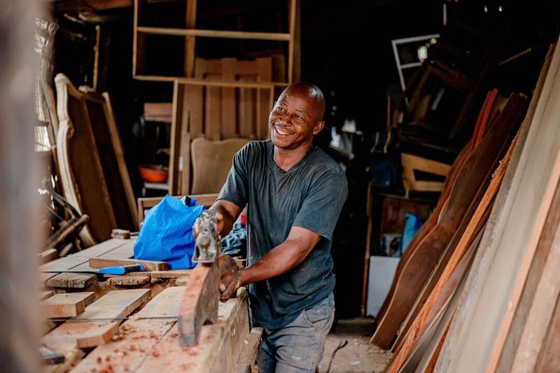 A carpenter working a board by hand in his own small workshop, surrounded by stacked timber.