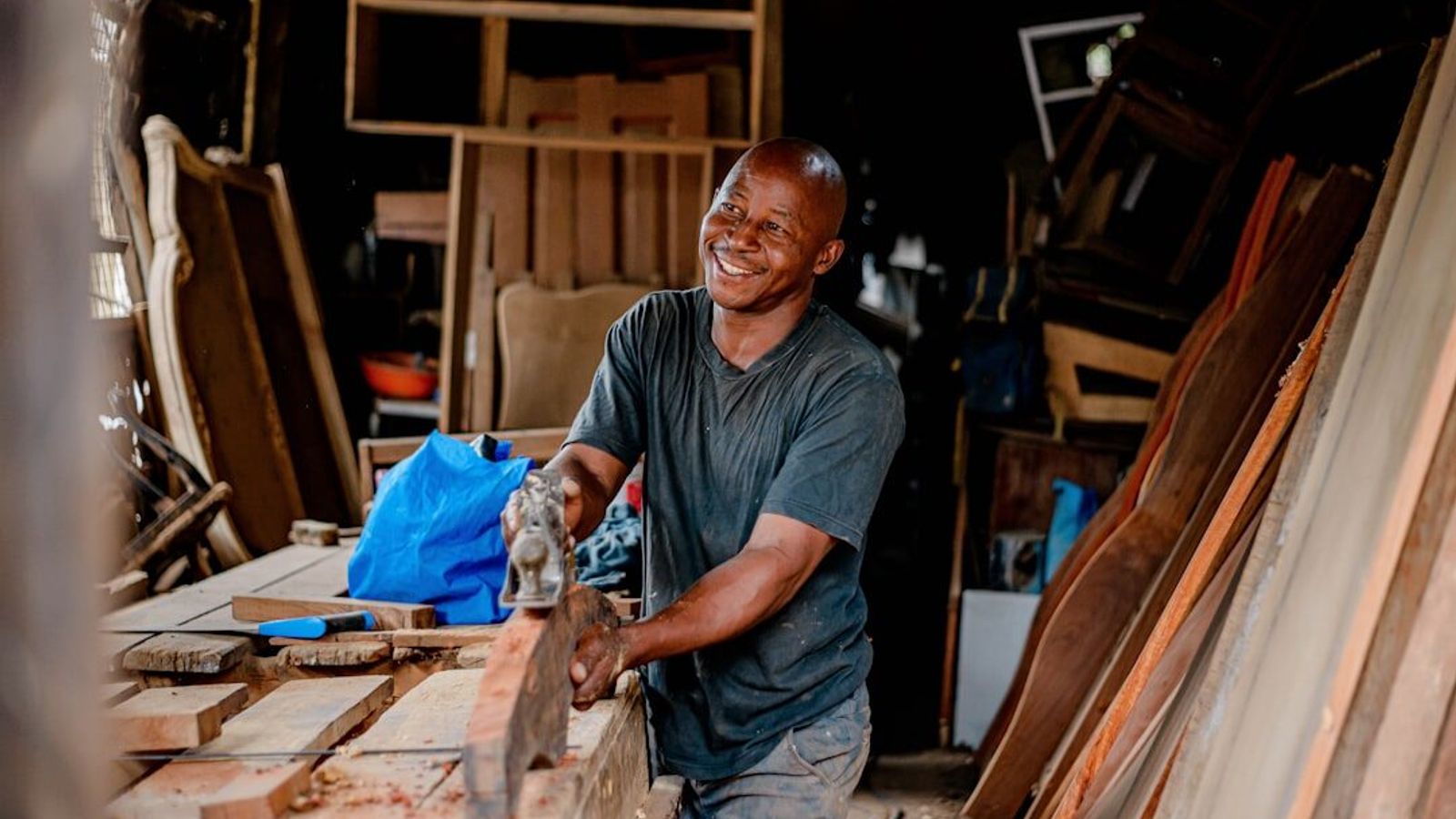 A carpenter working a board by hand in his own small workshop, surrounded by stacked timber.