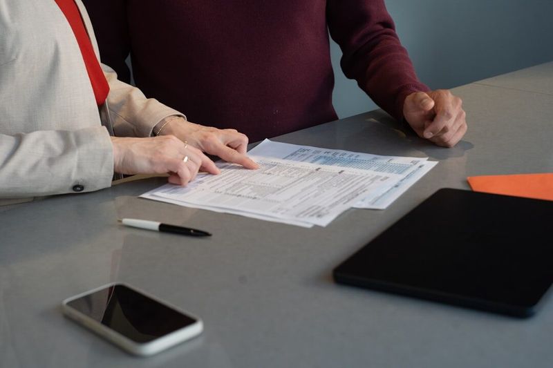 Two people at a conference table reviewing printed paperwork together, with a pen, phone and tablet on the surface between them.