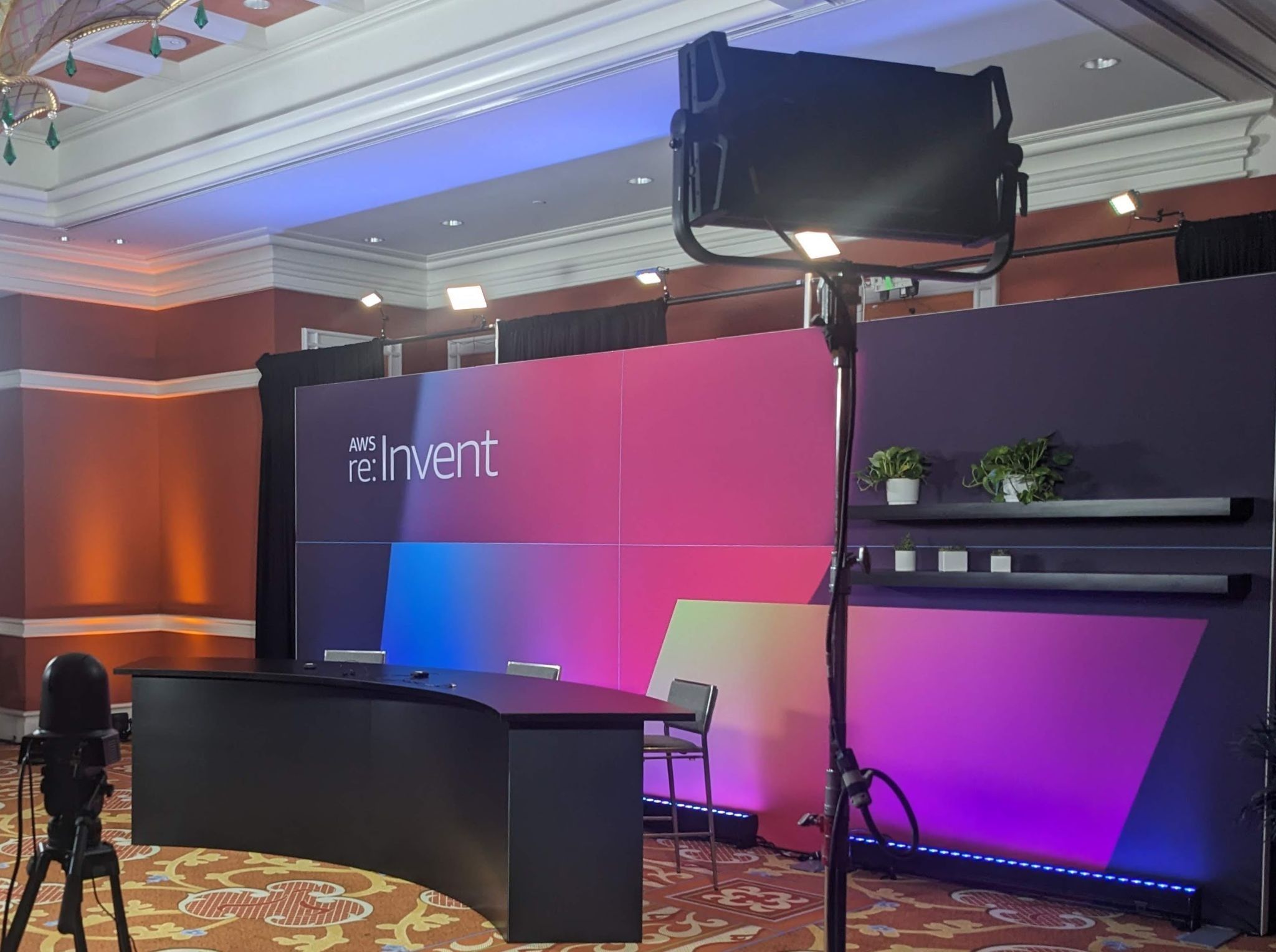 A curved desk with two chairs sits in front of a background that displays the re:Invent logo. Cameras and lights are pointed at the desk.