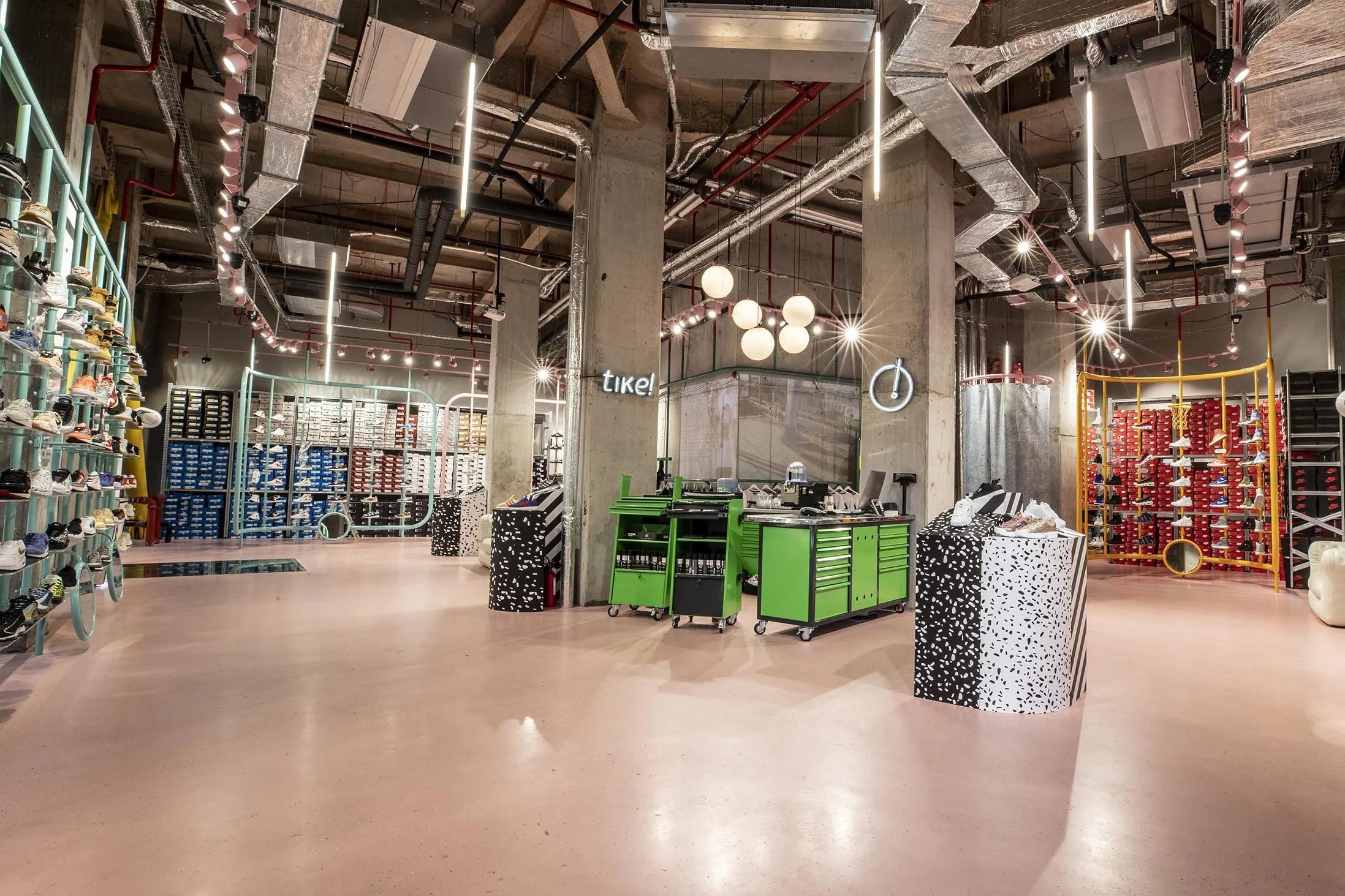 A modern, industrial-style shoe store with pink flooring, exposed concrete, teal and yellow sneaker displays, and green counters.