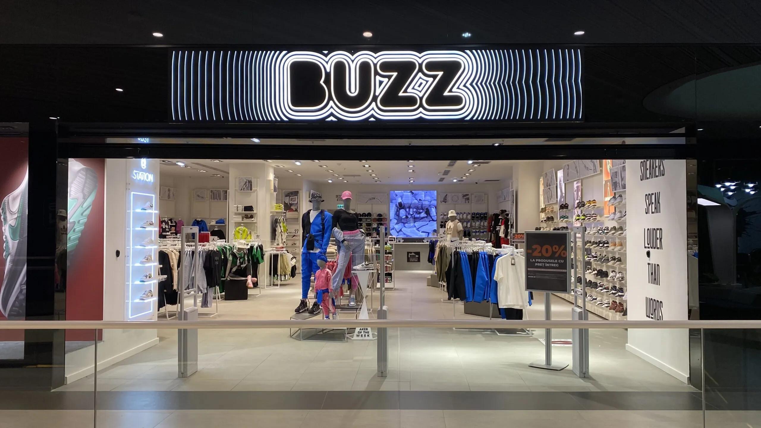 Storefront of a "BUZZ" clothing and shoe store, featuring an illuminated sign, mannequins, and merchandise.