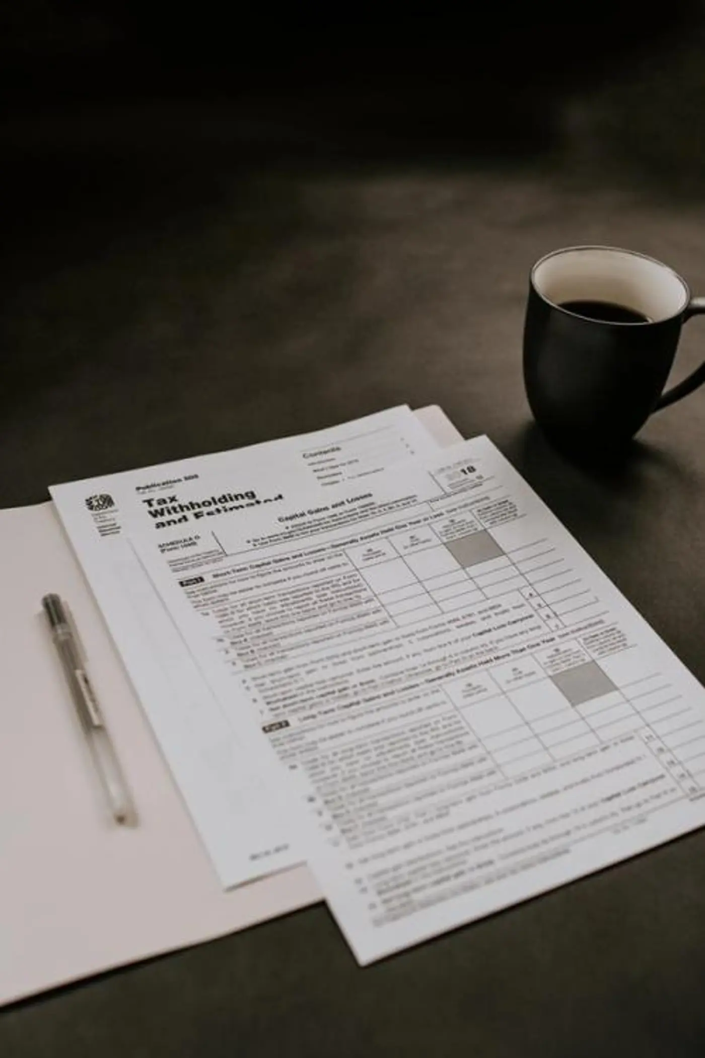 A stack of tax documents, including a U.S. tax form titled ‘Tax Withholding and Estimated Tax,’ is placed on a dark surface. A pen rests on a folder beside the papers. A black coffee mug filled with a dark liquid sits nearby.