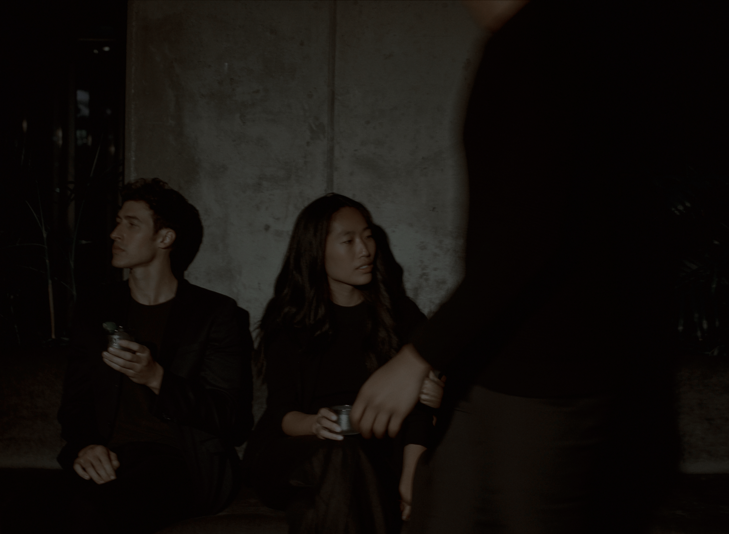 A young man and woman sit on a bench with drinks in hand while someone walks past