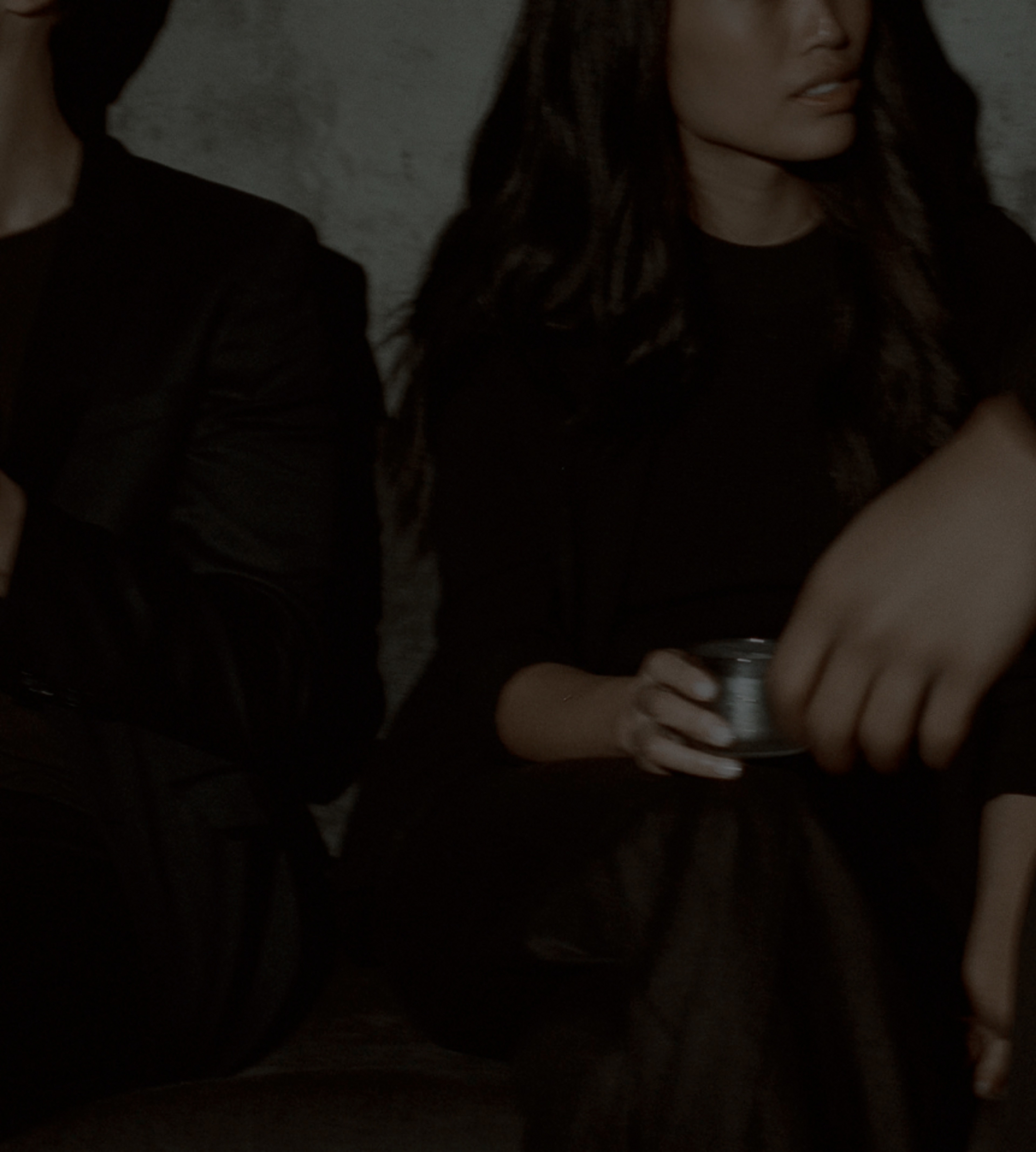 A young man and woman sit on a bench with drinks in hand while someone walks past
