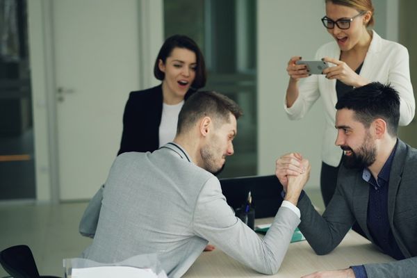 Colleagues arm wrestling while others watch and film.