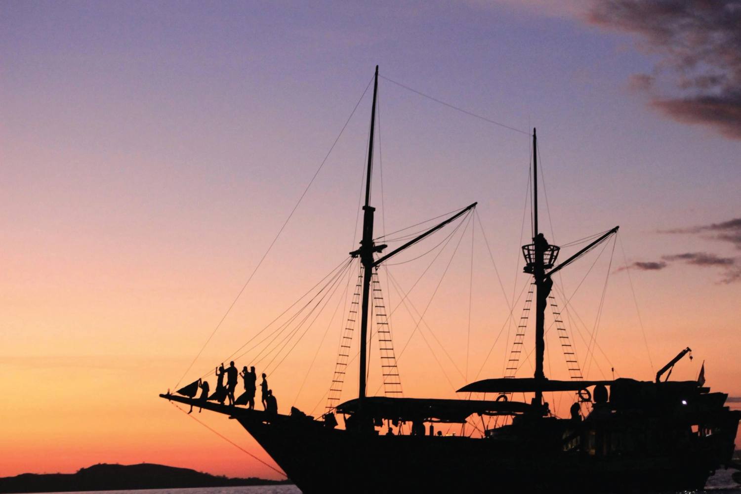 A traditional phinisi in silhouette at sunset, guests gathered on the bowsprit