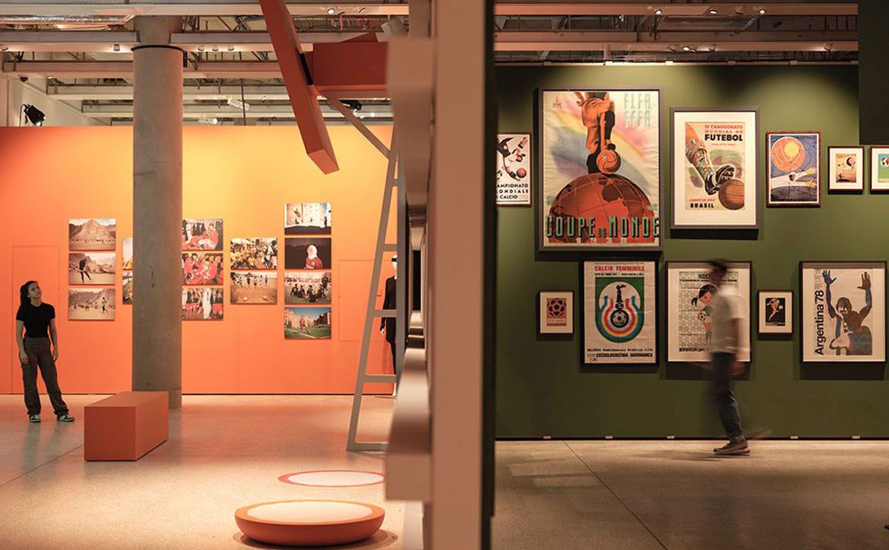 Two visitors walking around the exhibition, Football: Designing the Beautiful Game, looking at football posters, at the Design Museum. Exhibition Soundscape created by Coda to Coda